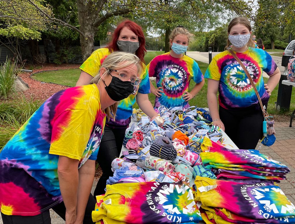 Group of women in tie-dye shirts and masks at outdoor event table.