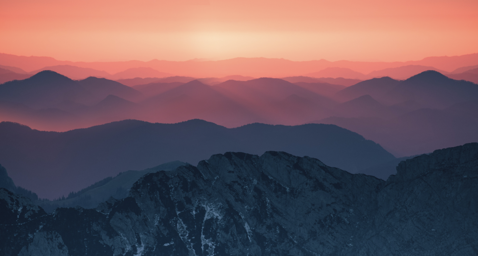 Mountain range at sunset with layered silhouettes and orange sky.